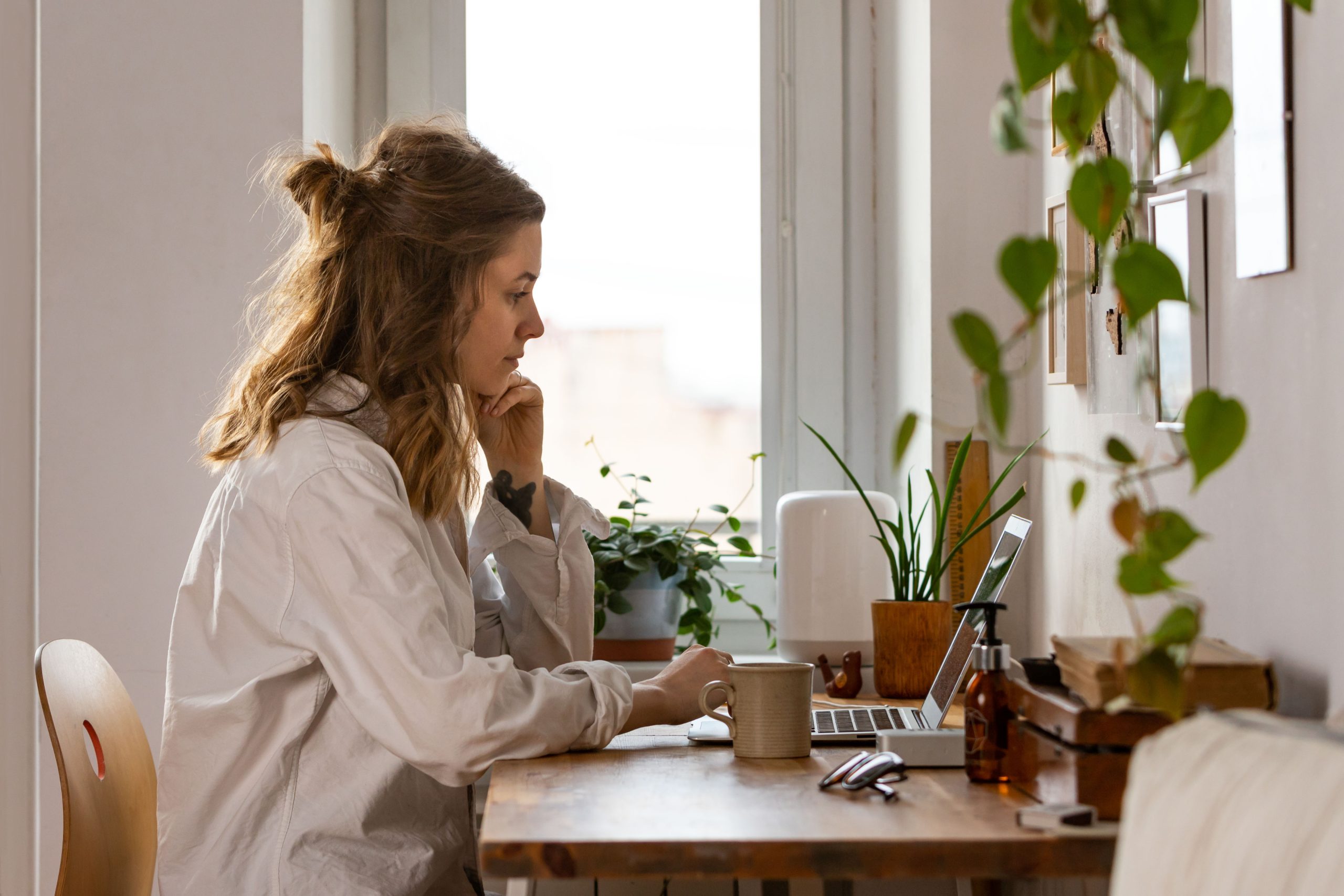 Woman sat at a desk next to a window with a laptop, coffee mug, and houseplants.