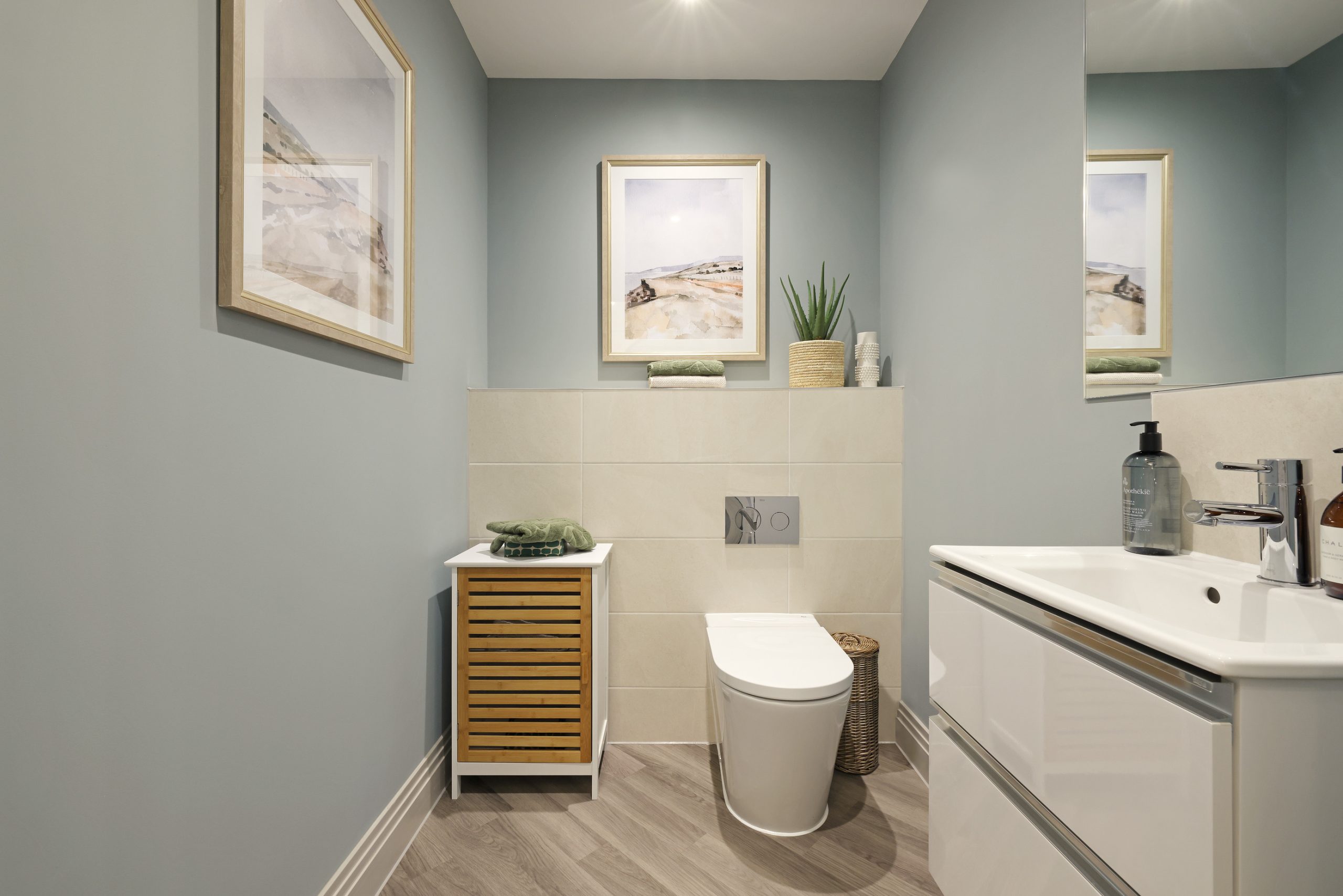 Modern bathroom with beige tiles, grey walls, framed art, and a large mirror mounted on the right wall above the sink.