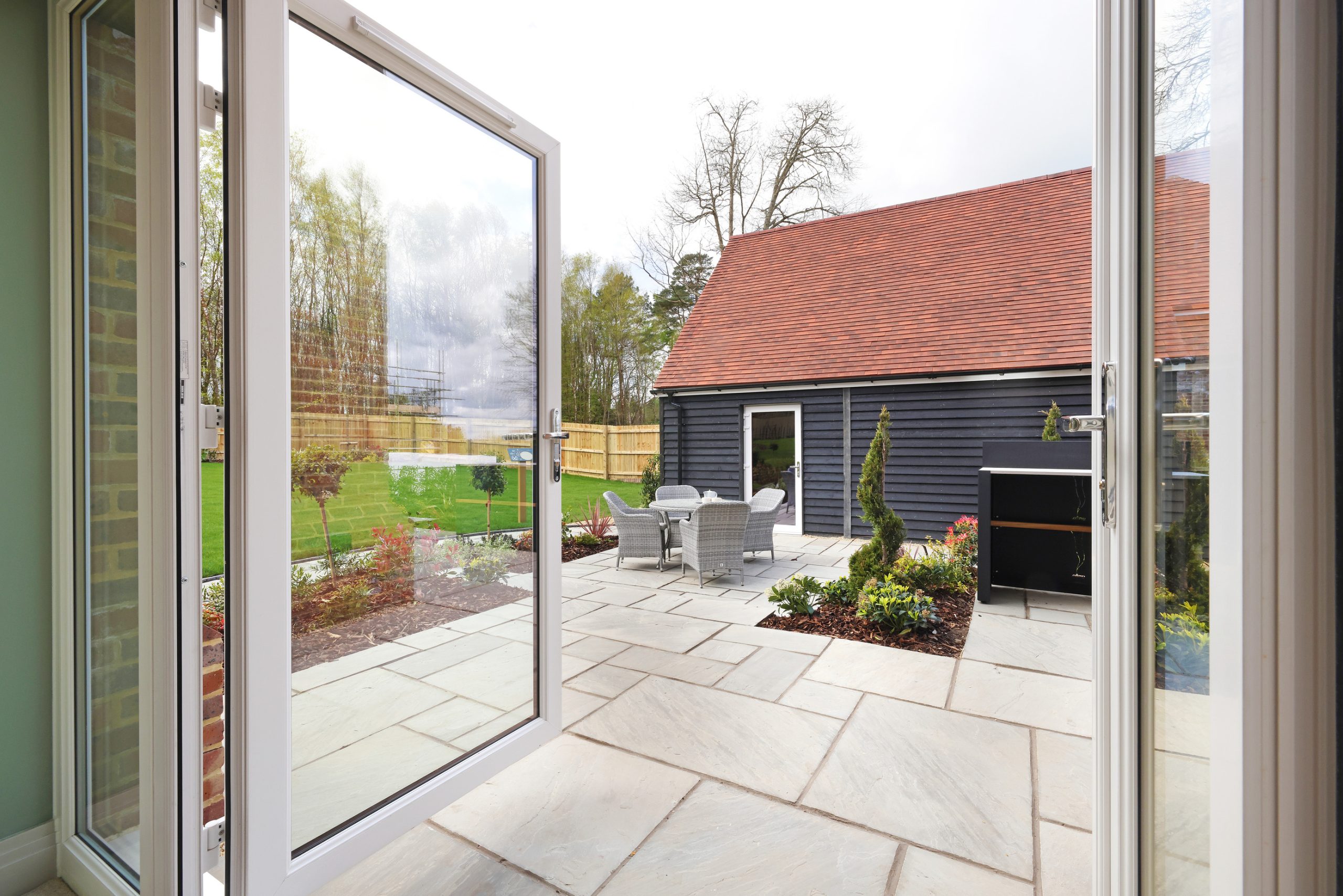 View from open door onto a patio with furniture and a garden leading to an outhouse building